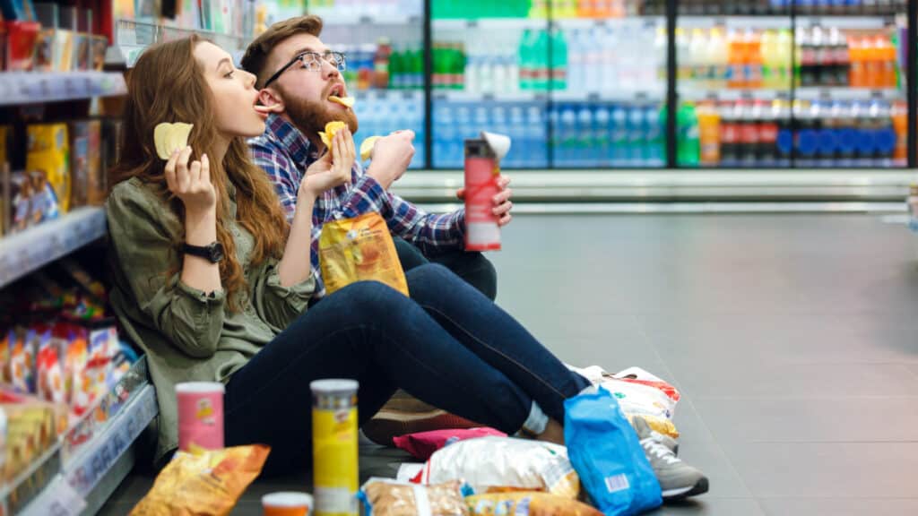 Man and woman sitting on floor of supermarket eating junk food. 