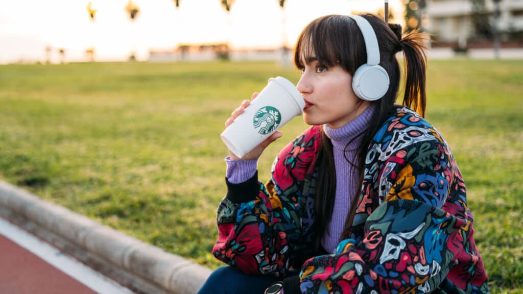 Young woman with sweater and headphones enjoying Starbucks coffee sitting outside.