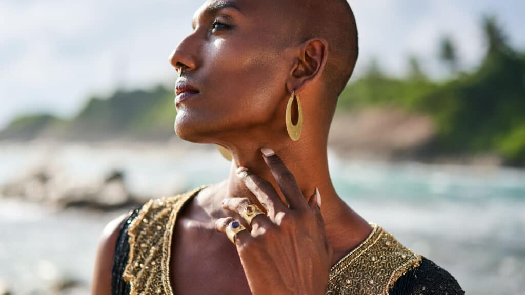 Trans sexual ethnic fashion model in long posh dress and accessories in elegant posture touches neck. Epatage gay black man in luxury jewelry poses, touches neck on scenic ocean beach. Pride, close up