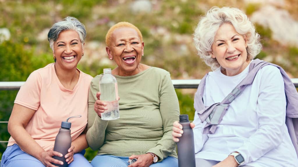 Three older women sitting on bench drinking water.