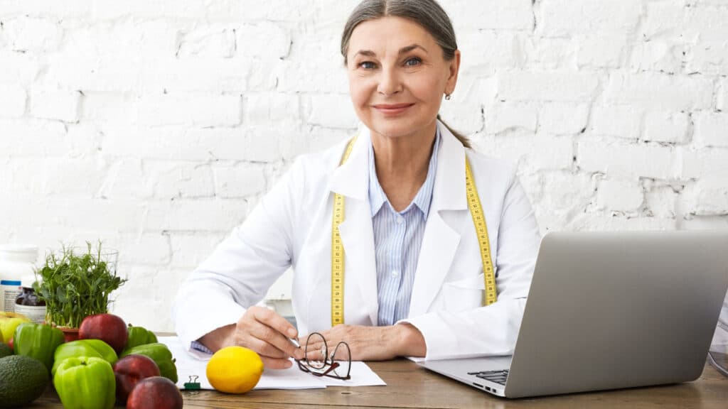 Woman in white coat with fruit and vegetables on table. Dietitian.
