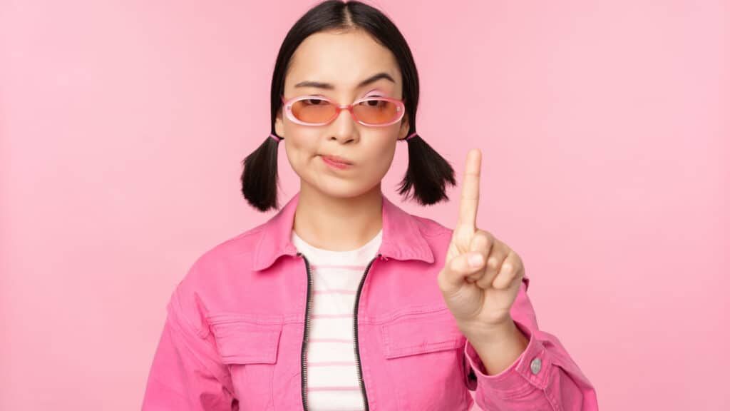 Asian woman in ponytails, wearing pink, saying no, wagging finger.