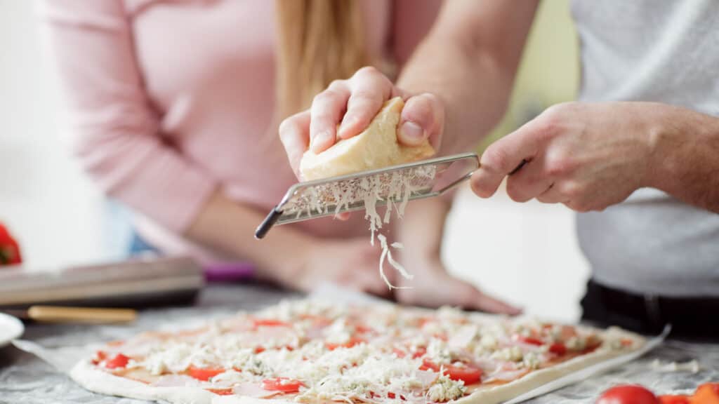 Hands grating cheese onto pizza. 
