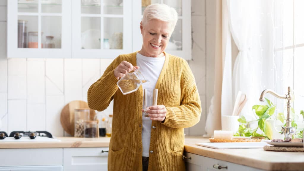Older woman pouring a glass of water to drink. Hydration.
