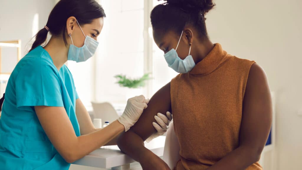 Young African American woman getting flu shot during seasonal vaccination campaign. Doctor or nurse in medical face mask cleans skin on patient's arm before injecting modern Covid 19 antiviral vaccine