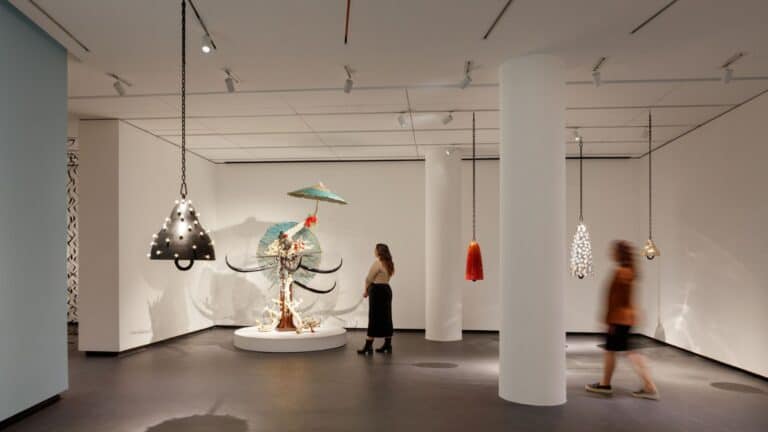 A modern museum gallery is photographed at a wide angle. Visitors observe large sculptures, several of which hang from the ceiling.