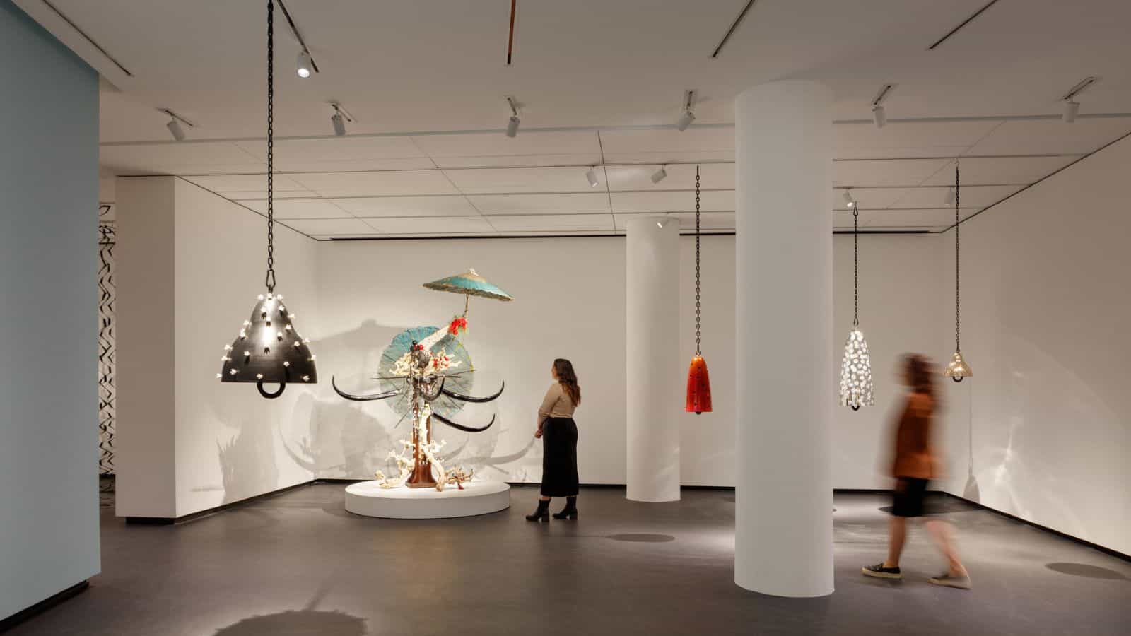 A modern museum gallery is photographed at a wide angle. Visitors observe large sculptures, several of which hang from the ceiling.