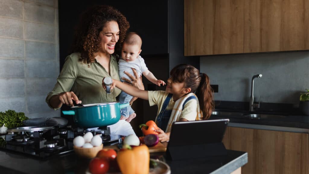 Young mother with baby and young daughter cooking in kitchen. 