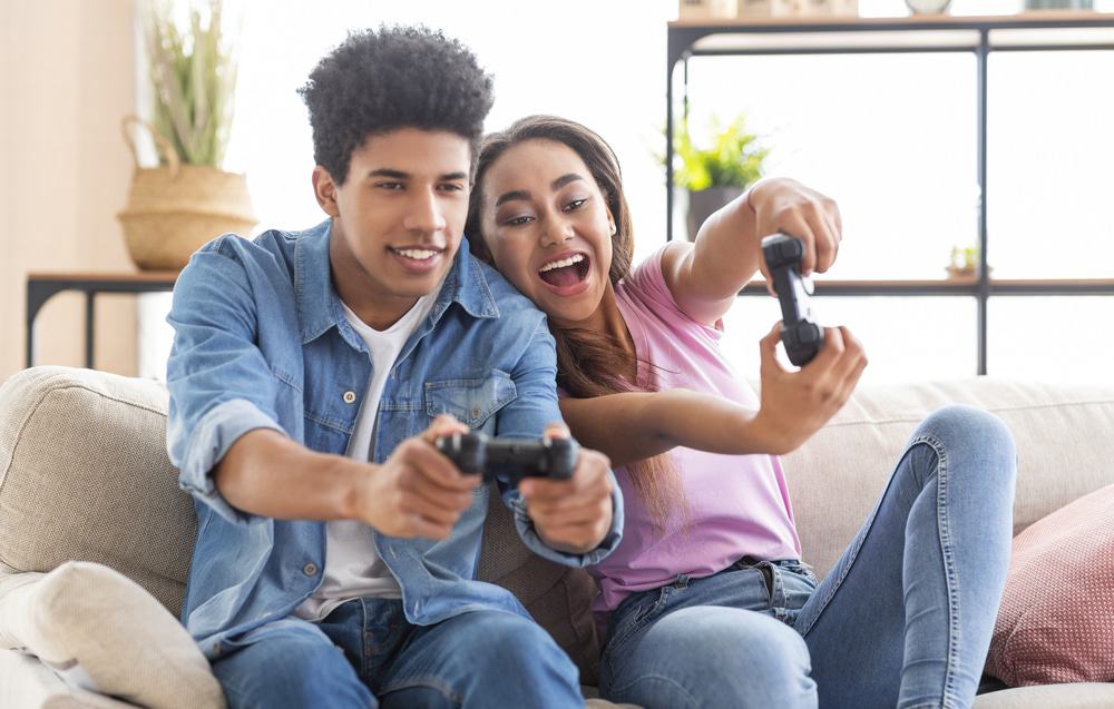 Happy black teenagers playing video game with control pad, holding joysticks in living room