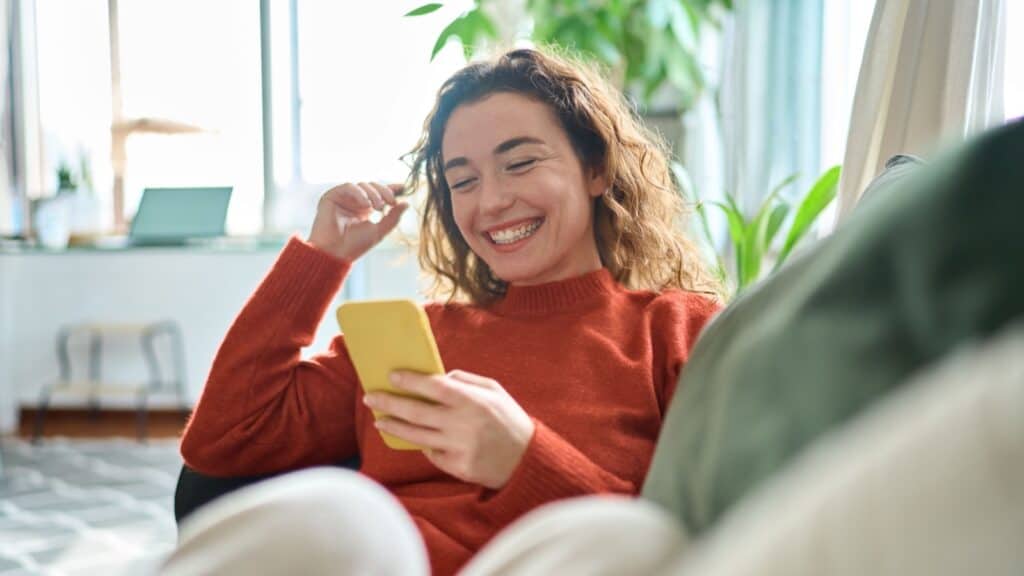 Happy relaxed young woman sitting on couch using cell phone, smiling lady laughing holding smartphone, looking at cellphone enjoying doing online ecommerce shopping in mobile apps or watching videos.