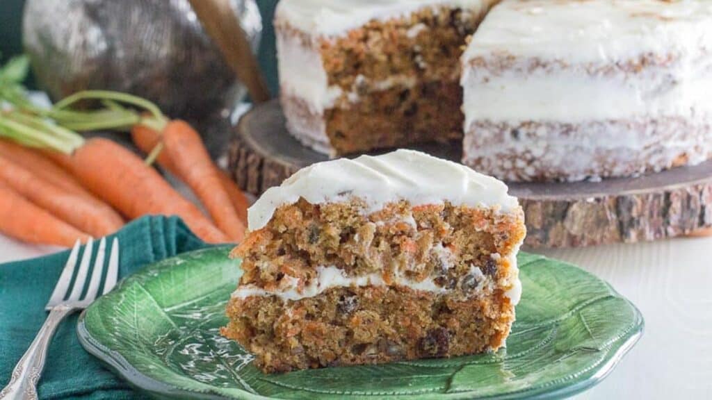 slice-of-low-FODMAP-carrot-cake-with-cream-cheese-frosting-on-green-plate-with-carrots-and-whole-cake-in-background.
