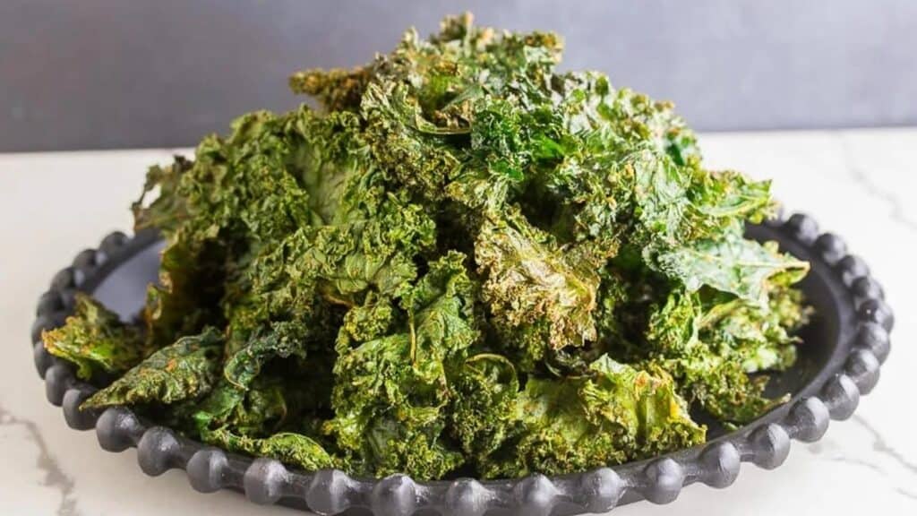 Roasted-kale-chips-piled-up-on-black-plate-white-surface-dark-background.