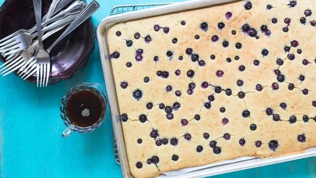 Sheet-Pan-Pancakes-with-Blueberries-in-pan-on-cooling-rack-against-aqua-background-maple-syrup-on-the-side-in-glass-pitcher.