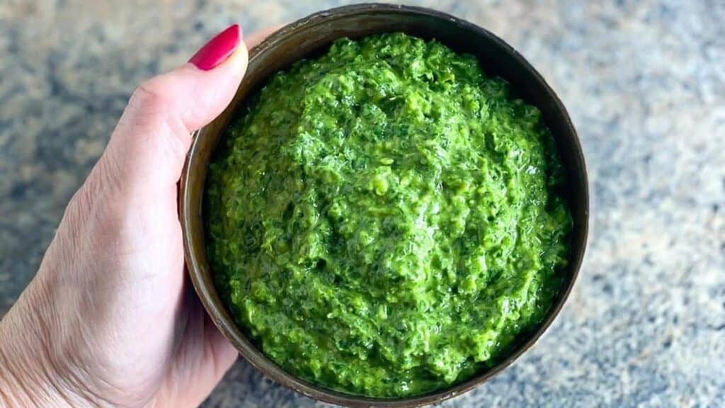 horizontal-image-of-vegan-low-FODMAP-kale-pesto-in-a-brown-bowl-held-by-womans-manicured-hand-over-stove-surface.