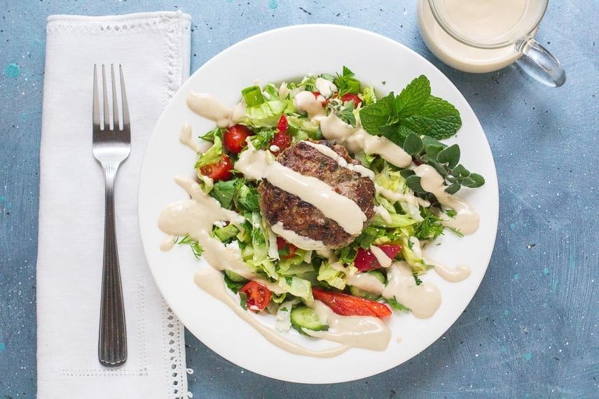 overhead-shot-of-low-FODMAP-Lamb-Burgers-with-Tahini-Sauce-on-salad-and-white-plate-linen-napkin-and-fork-alongside.
