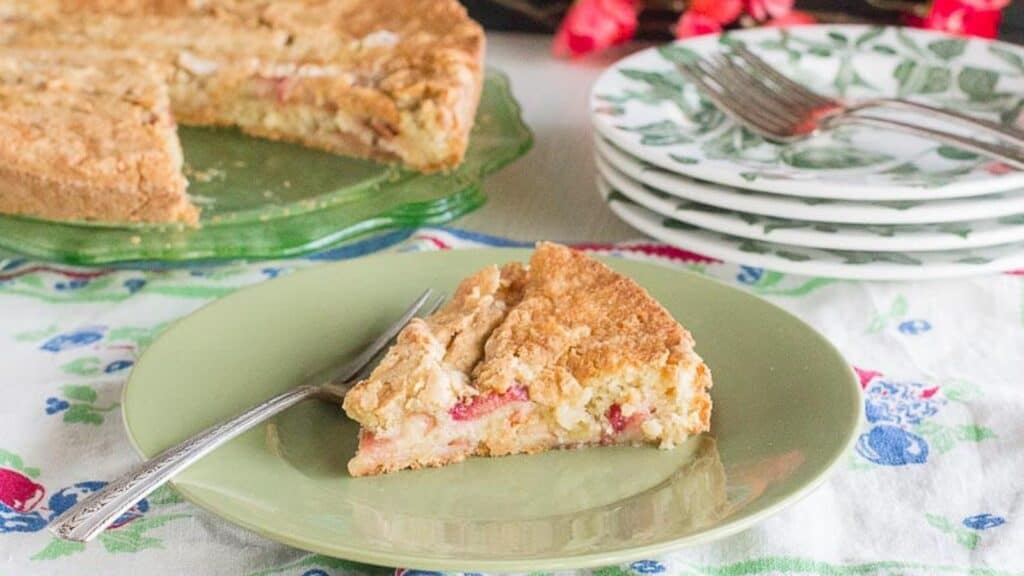rustic-rhubarb-cake-slice-on-green-plate-whole-cake-in-background-on-green-glass-plate.-Floral-linens-and-red-blossoms-in-background.