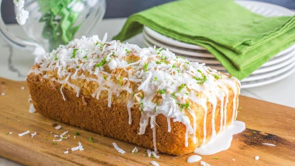 side-view-of-coconut-lime-bread-on-a-wooden-board-icing-dripping-down-white-plates-and-green-napkin-in-background.