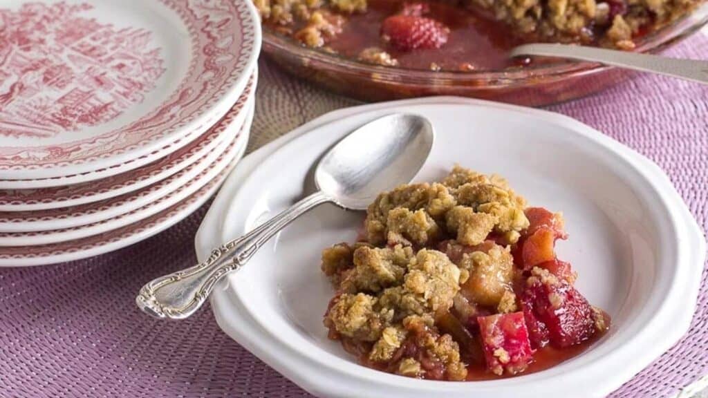 strawberry-rhubarb-crisp-on-a-white-plate-with-silver-spoon-stack-of-pink-plates-and-whole-crisp-in-background.