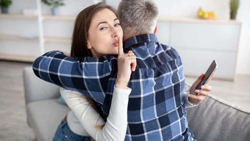 Beautiful mature woman hugging her husband, showing HUSH gesture, having secret affair, texting her lover on smartphone. Adultery, relationship issue, marriage cheating, hidden love concept