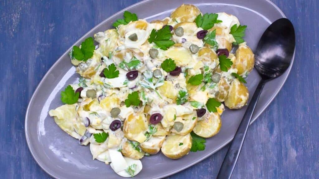 Tangy-Potato-Egg-Salad-with-Olives-Pickles-on-a-grey-oval-platter-against-a-blue-backdrop.