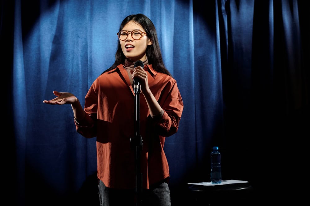 Young,Comedian,In,Eyeglasses,And,Brown,Shirt,Standing,On,Stage