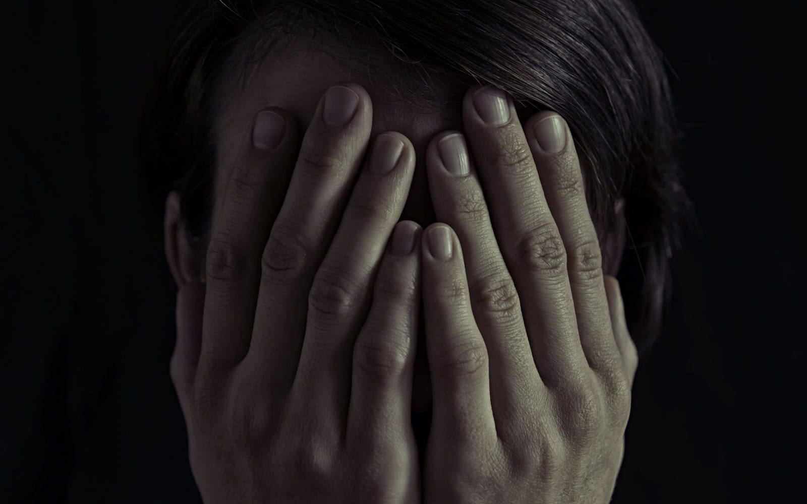 Concept of fear, domestic violence. Woman covers her face her hands. Dim light and black background , creates a dramatic mood of this image.
