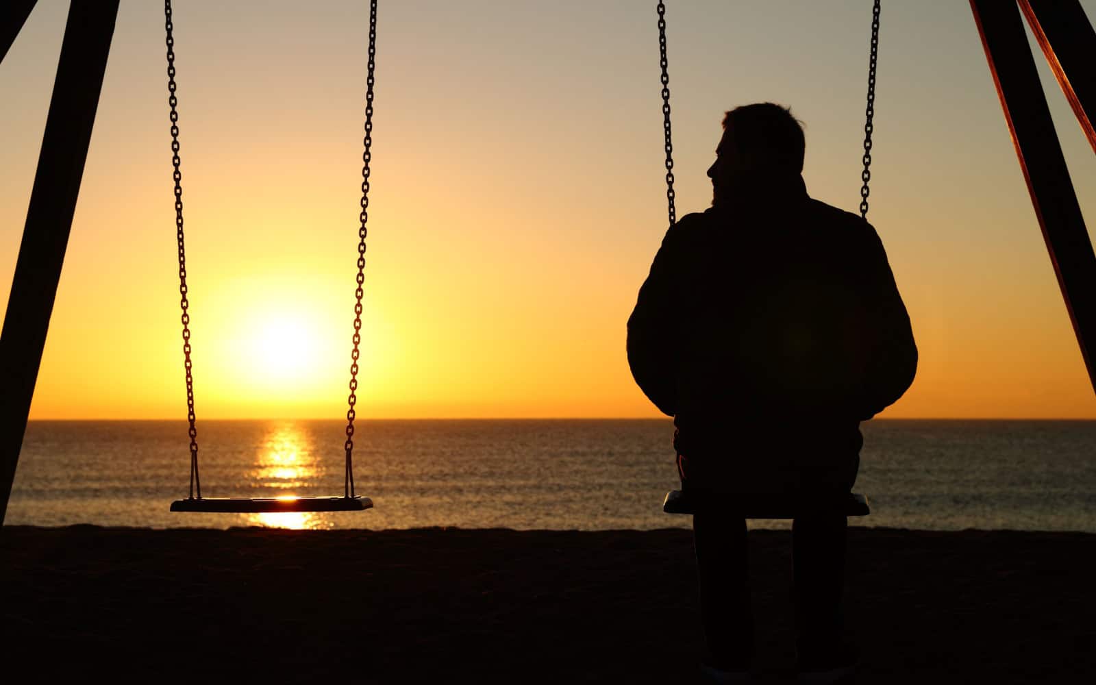 Back view backlighting silhouette of a man alone on a swing looking at empty seat at sunset on the beach in winter