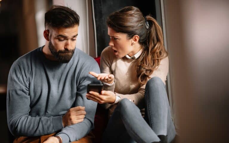 Angry couple or marriage fighting for a mobile phone at home. Jealous caucasian woman holding smart phone and showing message to his husband