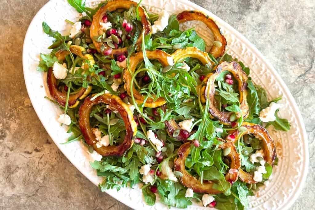 Roasted-Delicata-Squash-Arugula-Salad-with-Goat-Cheese-and-Pomegranate-on-a-white-oval-platter-and-stone-surface.