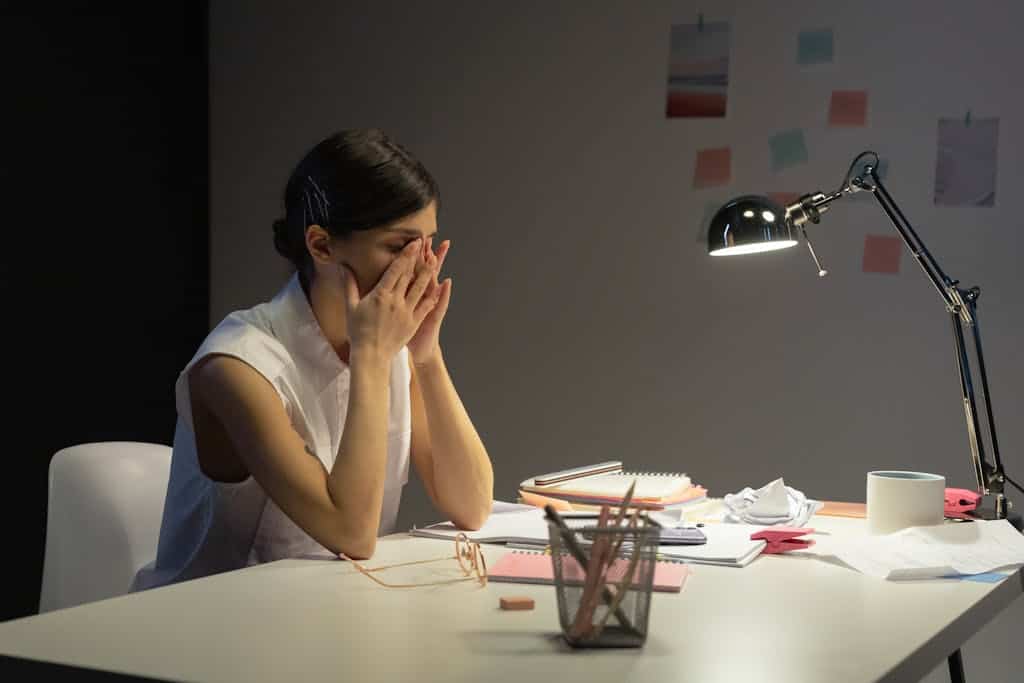 A stressed businesswoman rubs her eyes at a cluttered desk, overwhelmed by work.