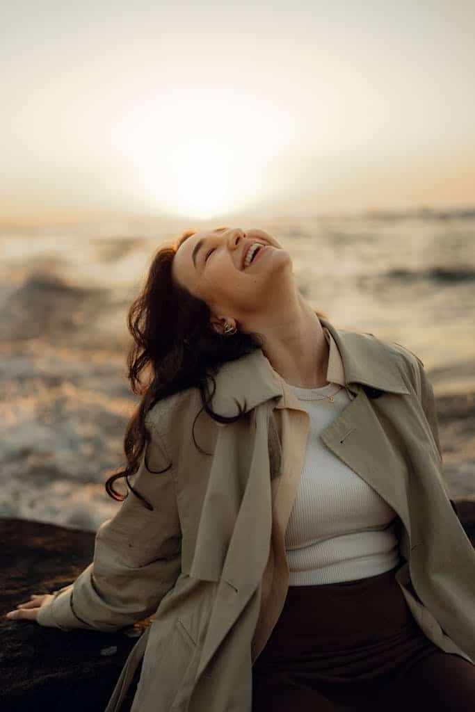 A woman in a trench coat enjoys a sunset by the ocean, exuding joy and serenity.