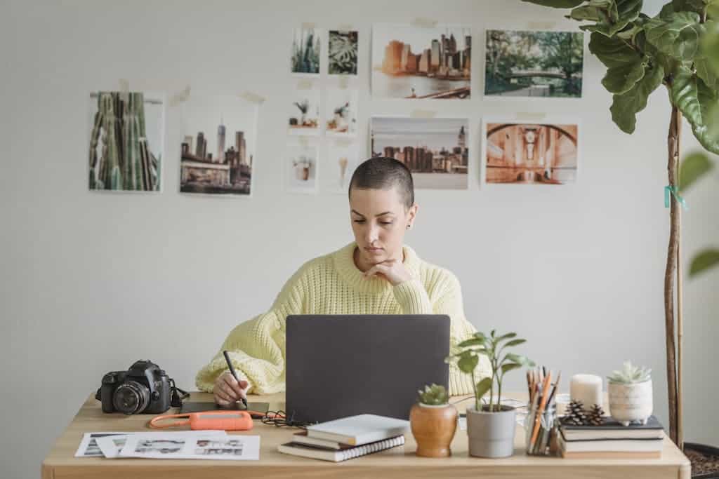 Female graphic designer working on a laptop with a stylus in a cozy home office filled with plants and art.