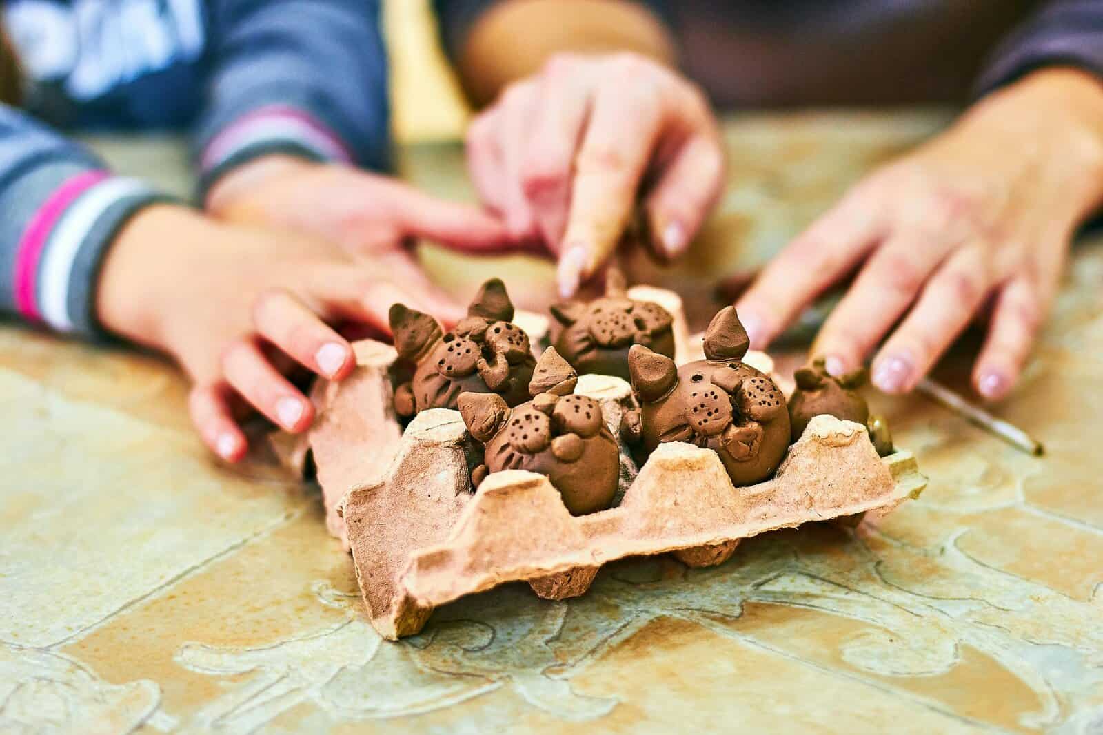Hands of children crafting clay animals in an egg carton, showcasing creativity.