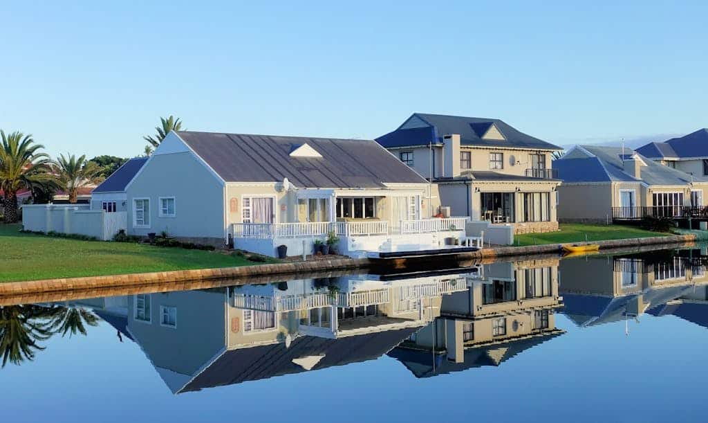 Luxury modern homes reflecting on a peaceful, clear lake during daylight, showcasing architectural beauty.