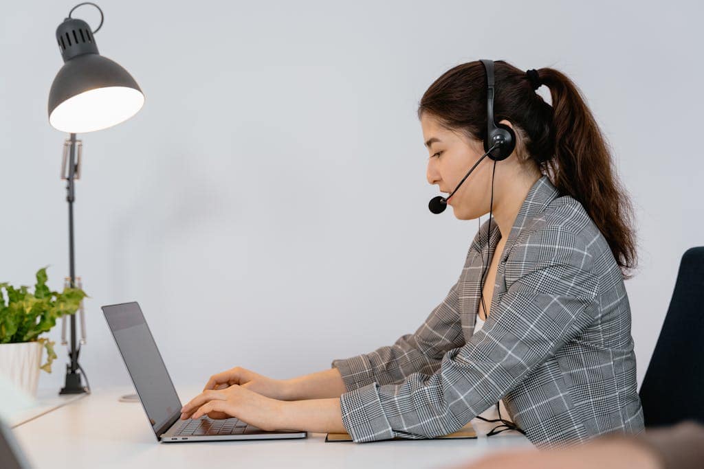 Professional woman in office with headset typing on laptop, offering customer support.