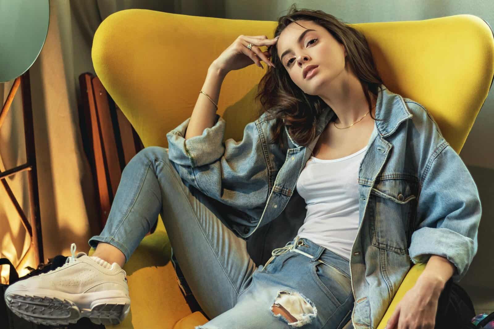 Stylish woman in denim poses on a modern yellow chair indoors.