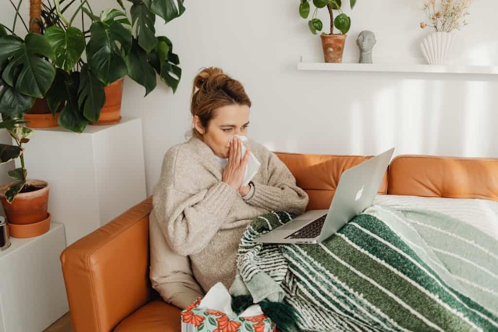 Woman in cozy sweater using laptop on sofa, surrounded by indoor plants, while blowing nose.