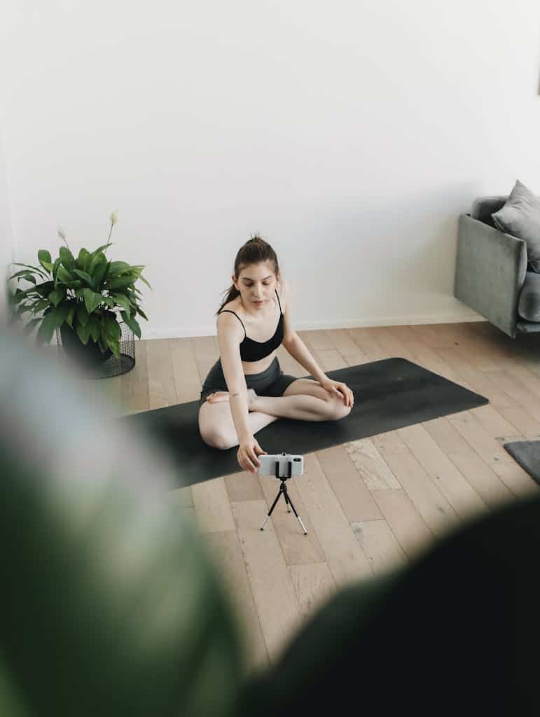 Woman in yoga pose on mat, filming herself with a smartphone indoors.