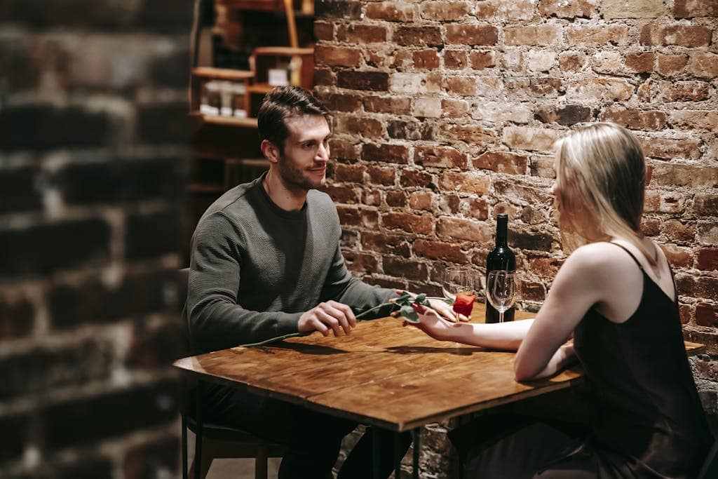 Loving boyfriend giving red rose to girlfriend while sitting near brick wall at wooden table during celebration of event in restaurant