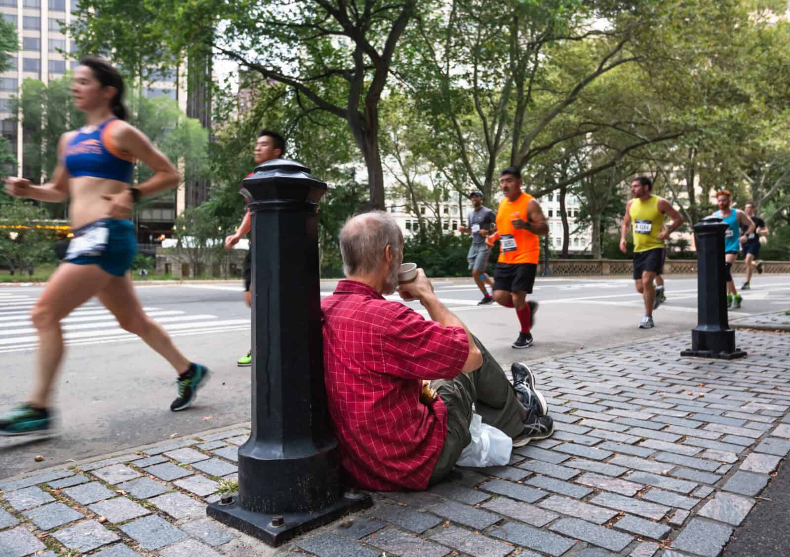 New York, USA - Sep 18, 2016: Manhattan street scene. An elderly man with a cup of coffee is sitting on the sidewalk near the central park in New York, watching people running along the street
