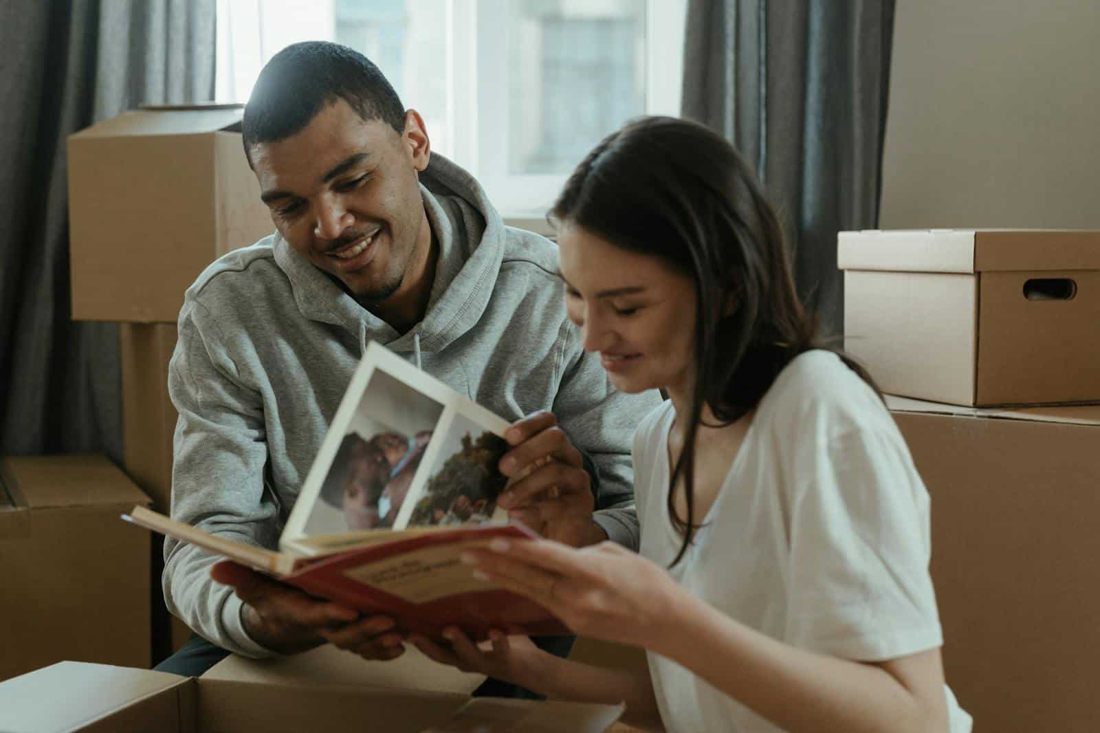 A happy couple reminisces over a photo album while unpacking boxes in their new home.