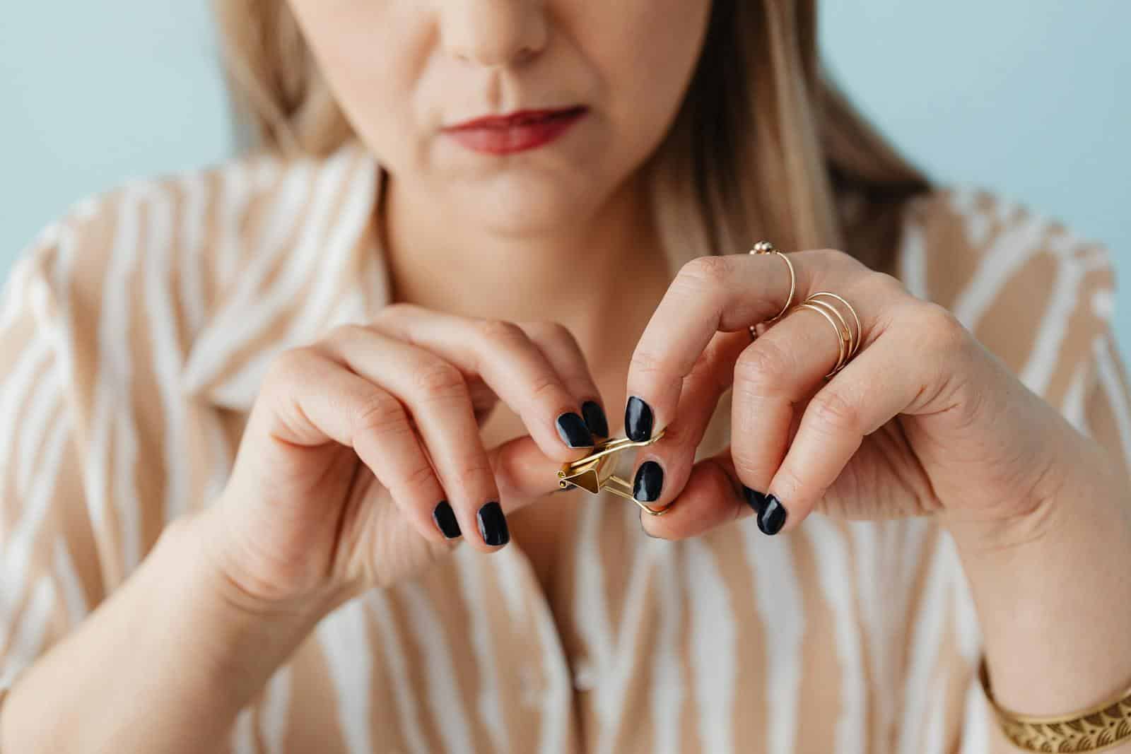 Close-up of a woman's hands fidgeting with a gold paperclip, showcasing painted nails and jewelry.