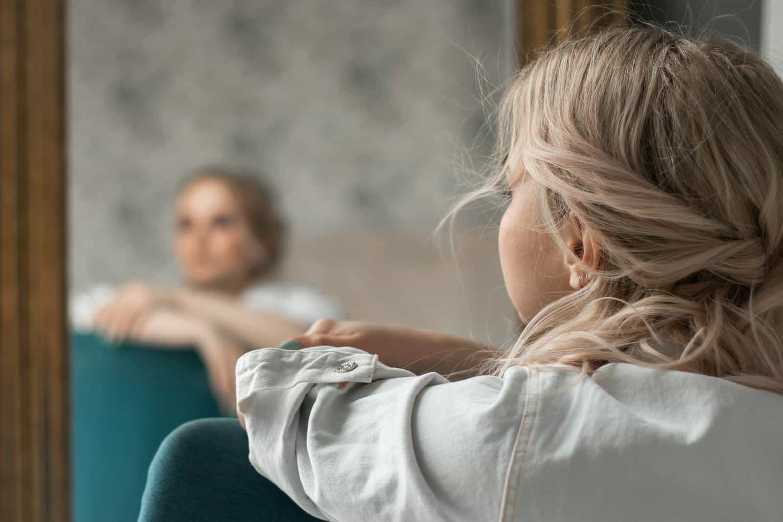 A contemplative woman with blonde hair gazes into a mirror, capturing a reflective indoor scene.