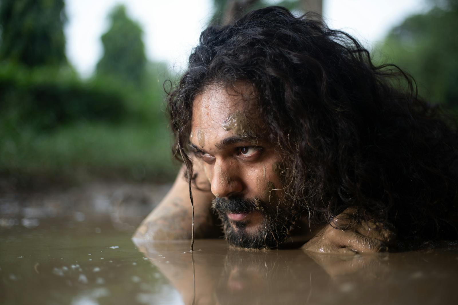 Bearded man with long hair in mud, focusing on survival and resilience.