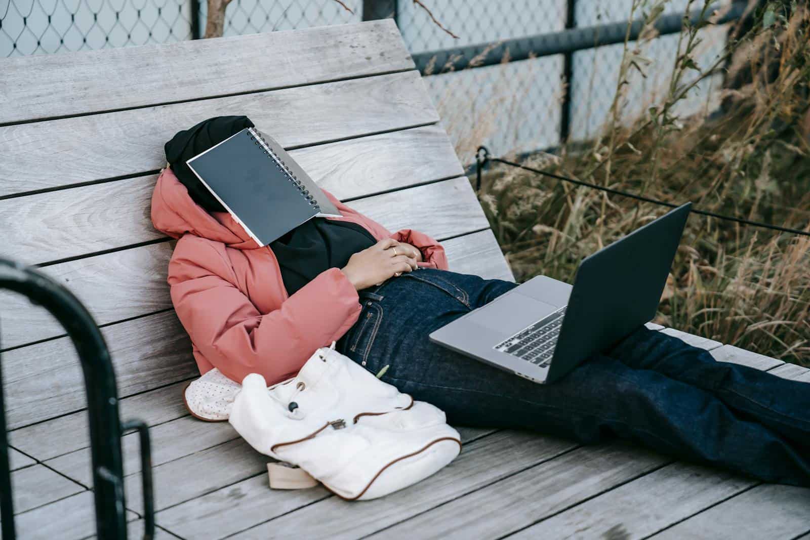 Tired female student in casual clothes and with laptop lying on wooden bench in city garden with notebook covering face