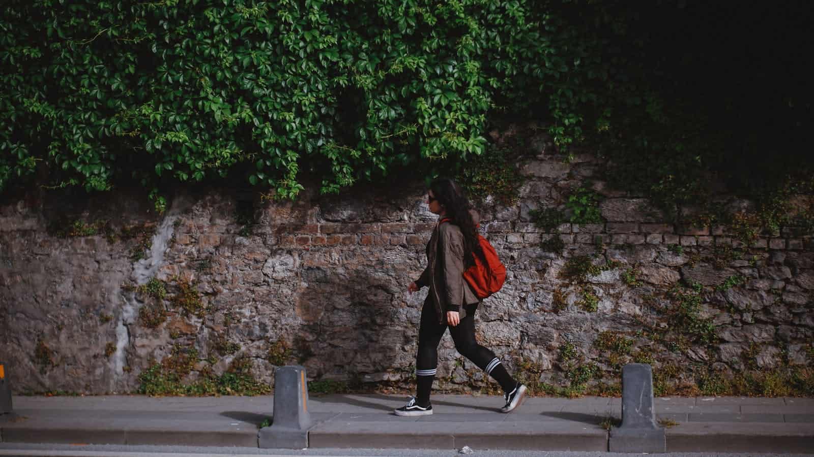 Woman walking with red backpack beside ivy-covered stone wall on urban sidewalk.