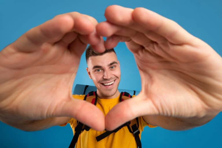 Happy male traveler with backpack making heart with his hands over blue studio background. Handsome camper with tourist equipment expressing his love. Summer holiday travel concept