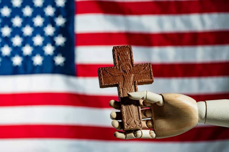 Wooden hand holding Christian cross in front of US flag. Mannequin hand holding a carved wooden cross on American flag background. Concept of religion faith and Christian values in the United States