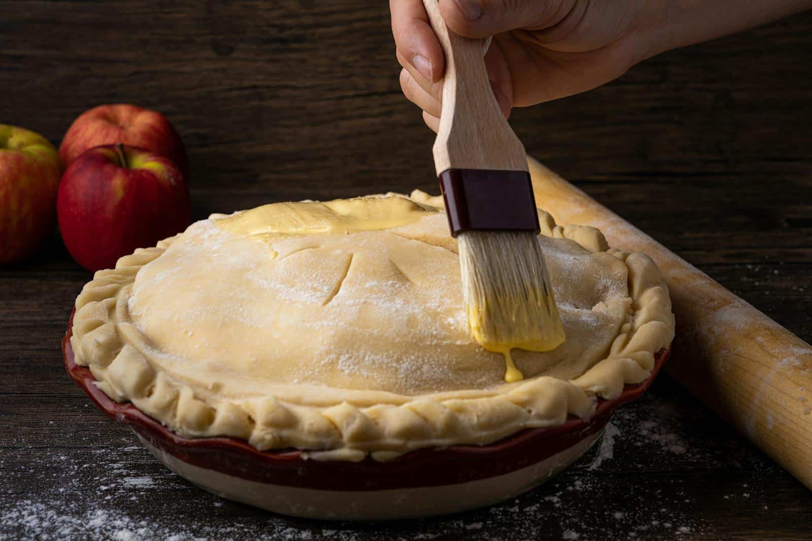 A close-up view of a homemade apple pie being prepared with an egg wash on a wooden surface.