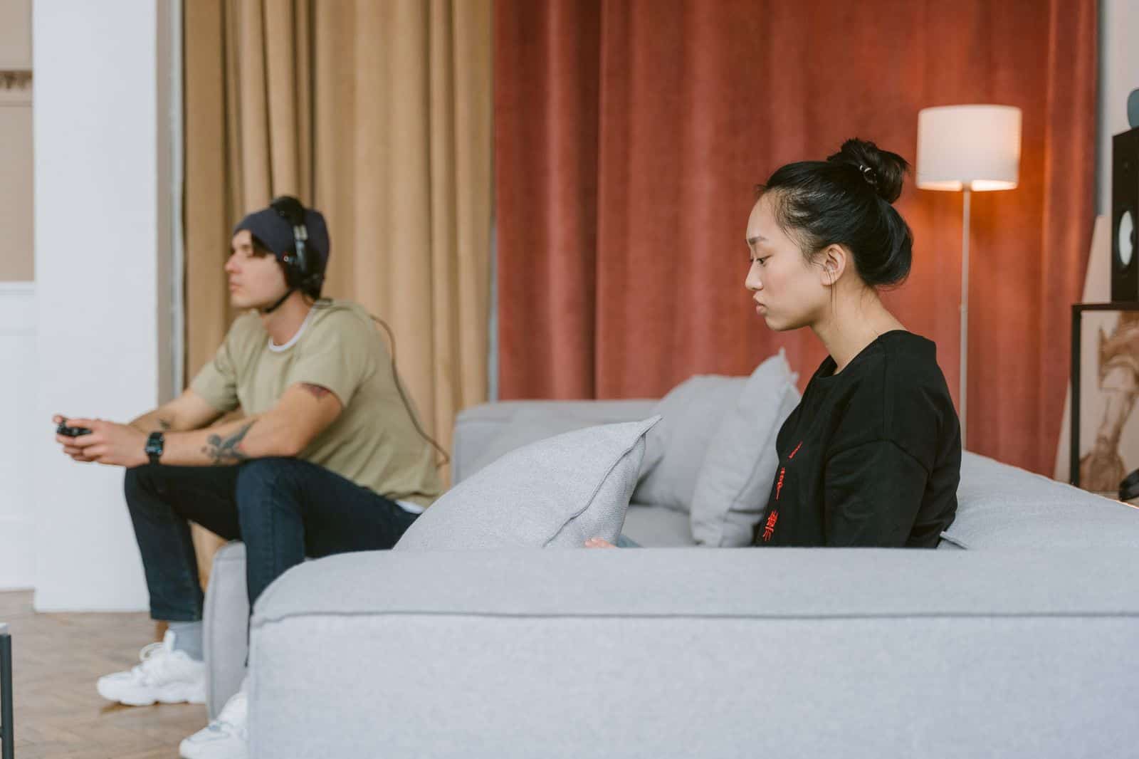 A couple sits on a couch, illustrating tension as one plays video games and the other appears distant. couple watching partner play video game